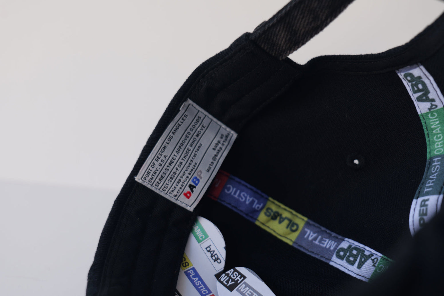 Interior view of the bABP B-SIDE black washed cotton hat showing the "Port of Design: Los Angeles" heritage label, branded recycling-themed seam tapes, and internal logo embroidery