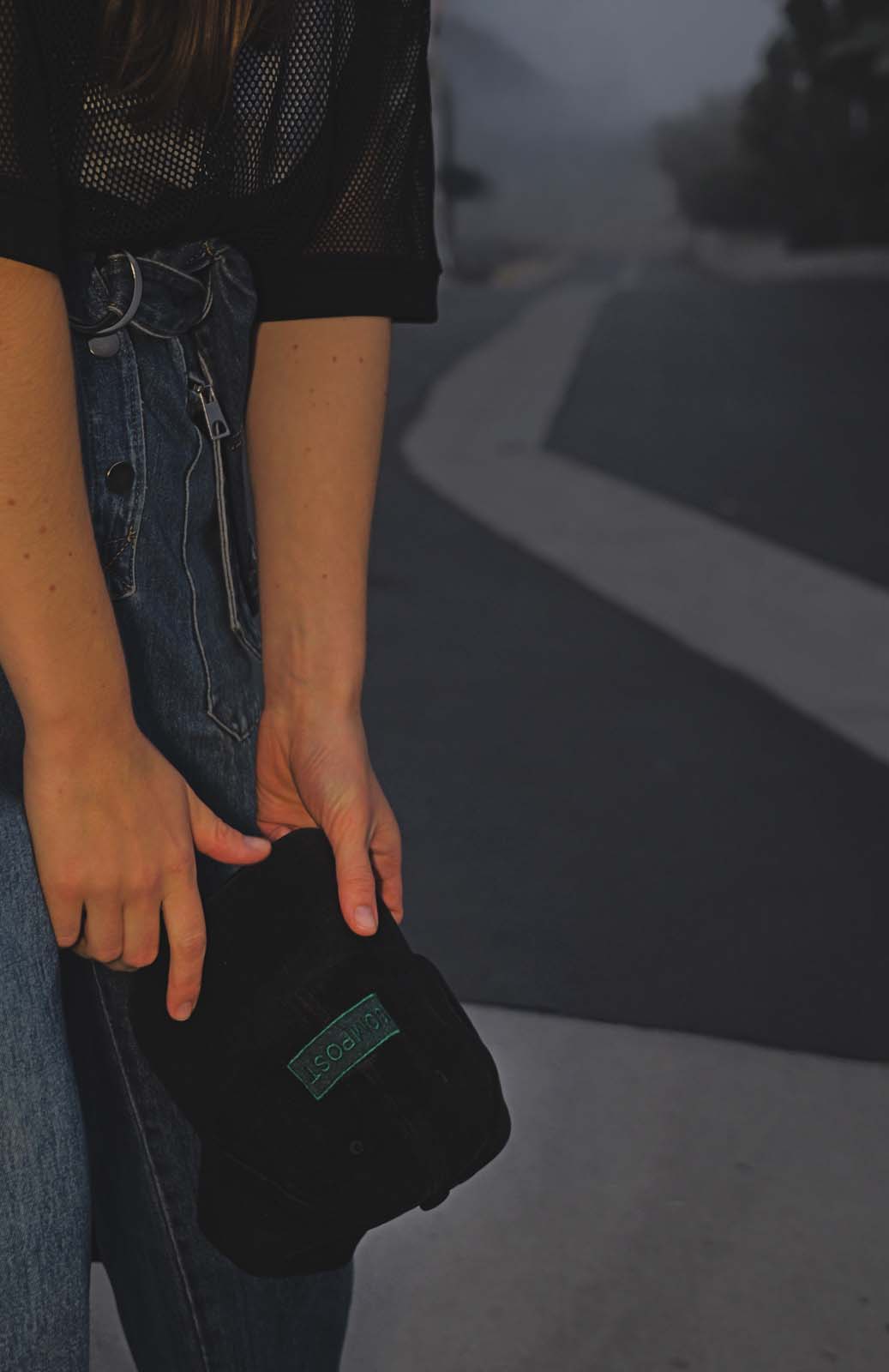 Model in a denim jumpsuit holding the bABP B-SIDE "COMPOST" black washed cotton hat with a green logo patch against a moody foggy road background