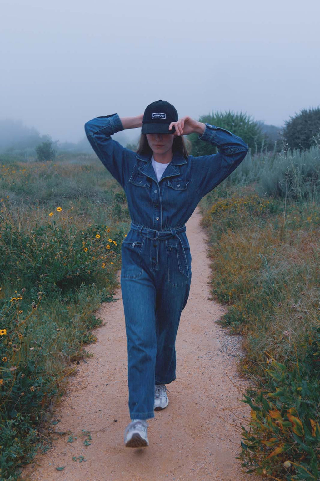 Model in a blue denim jumpsuit walking towards the camera on a dirt path in a foggy field wearing the bABP B-SIDE "COMPOST" black washed cotton hat with a white logo patch